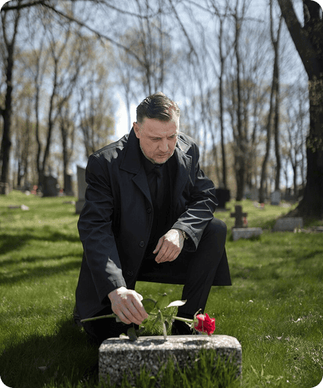 A man in a dark coat kneels in a cemetery, placing a red rose on a gravestone. Trees with bare branches and several other graves are visible in the background on a sunny day.